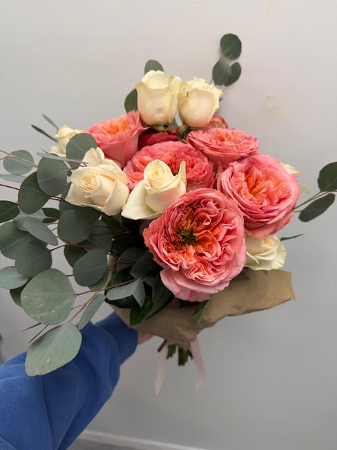 A hand holding a floral bouquet featuring large, ruffled pink and orange ranunculus flowers, interspersed with small, creamy white roses and sprigs of eucalyptus leaves with round, muted green foliage