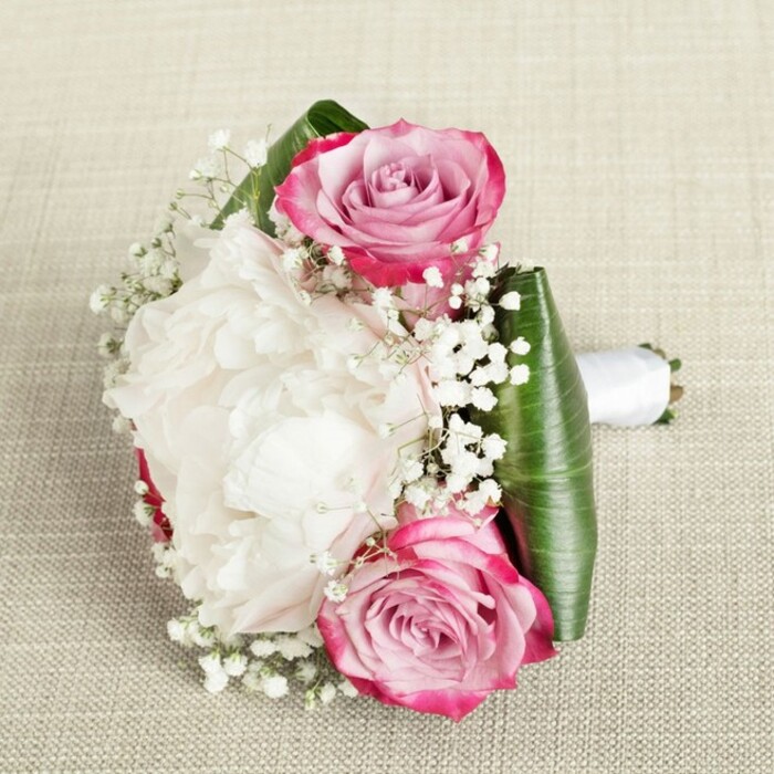 In the serene glow of North Canonbury, this elegant floral arrangement rests atop a textured, natural-fiber fabric, the sort one might find at the edge of New River Walk. The bouquet's composition is sophisticated and compact-two open pink roses, each suffused with shifting tones from vivid fuchsia to soft blush and just a hint of lavender, frame a luxurious, white peony in full bloom. The peony's tightly layered, ruffled petals create a sense of comforting abundance at the heart of the design, their creamy white hue enriched by gentle, ambient light. Delicate sprays of baby's breath thread throughout, filling spaces with their misty white clusters and infusing the bouquet with an airy, almost fairytale quality reminiscent of hidden Canonbury gardens. The base is wrapped with wide, deep emerald green leaves, possibly ti or aspidistra, expertly folded to cradle the arrangement and lend structure. Stems are neatly bound with white satin ribbon, its sheen catching the light and suggesting a touch of ceremony. The harmonious layering of blooms and foliage, together with the soft, tactile surface beneath, invites thoughts of quiet elegance and celebration. Whether intended for a local wedding, an anniversary, or to grace a family gathering near Canonbury's leafy lanes, this piece exudes both artistry and heartfelt emotion, representing the craft and warmth of local florists.