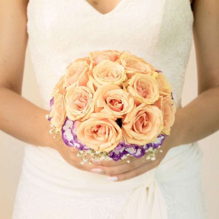 A detailed, chest-level photograph captures a bride holding her bouquet in the gentle light of what could be a Canonbury church vestibule or a bright townhouse hallway. The focus is on her hands and midsection: she wears a timeless white or ivory gown with fine lace detailing across the bodice and a soft V-neckline, the fabric gathered smoothly at the waist before flowing down in subtle folds. Her fingers encircle a round bridal bouquet that appears both structured and romantic, the roses arranged in a classic, domed shape. These roses, in shades of soft peach, pale apricot, and blush pink, dominate the upper portion of the arrangement. Each bloom is full and tightly layered, the petals curling inward in velvety spirals that speak of freshness and careful handling, as if the flowers have just been conditioned and arranged in a nearby Canonbury studio. Beneath this warm, pastel cluster runs a defined band of deep purple flowers edged gracefully in white-likely lisianthus-creating a ruffled border that frames the roses and introduces a rich, cool contrast. Around the base and between some of the blooms, fine sprays of tiny white flowers, reminiscent of baby's breath, add a light, cloud-like texture, softening the transition between the roses and their darker collar. The bride's nails are neatly manicured in a pale, understated shade, with a glimpse of delicate glitter on one thumb that mirrors the quiet glamour of the bouquet. The image conveys a feeling of poised anticipation, capturing the artistry of a local florist who understands how to pair classic roses with distinctive accent blooms for weddings held in Canonbury's leafy streets, intimate gardens, and historic venues.