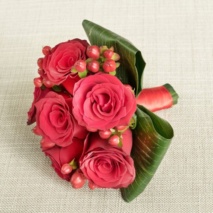 A carefully composed bouquet of deep red roses and berries rests on a canvas of light beige, subtly textured fabric, the neutral background chosen to let every nuance of red and green stand out. At the heart of the arrangement, around six large roses sit close together, each bloom in perfect condition. Their petals are dense and velvety, spiralling inward with an almost architectural precision, moving from near-burgundy shadows in the centre to glowing scarlet at the outer curves. Threaded among these roses are clusters of small, round Hypericum berries, ranging from soft, flushed red to deeper crimson, each berry perched on slender green stems that lend an airy, intricate texture between the fuller flowers. Two or three broad, dark green leaves sweep gracefully around one side of the bouquet, their edges gently curled. The leaves are glossy and slightly ribbed, creating a strong, sculptural frame that contrasts with the softness of the petals and the compact beads of the berries. The stems are neatly bound with a red satin ribbon wrapped tightly, adding a refined flourish that matches the roses' intensity. Light falls evenly across the scene, revealing every fold of fabric, petal, and leaf without harshness, conveying a tranquil, intimate atmosphere. This arrangement feels distinctly romantic-ideal for an anniversary, a special date night, or a discreet wedding posy in Canonbury, perhaps carried along St Paul's Road on the way to a ceremony. It captures the timeless symbolism of red roses-devotion, passion, and heartfelt affection-while the berries add a modern, playful touch that speaks of joy and celebration.