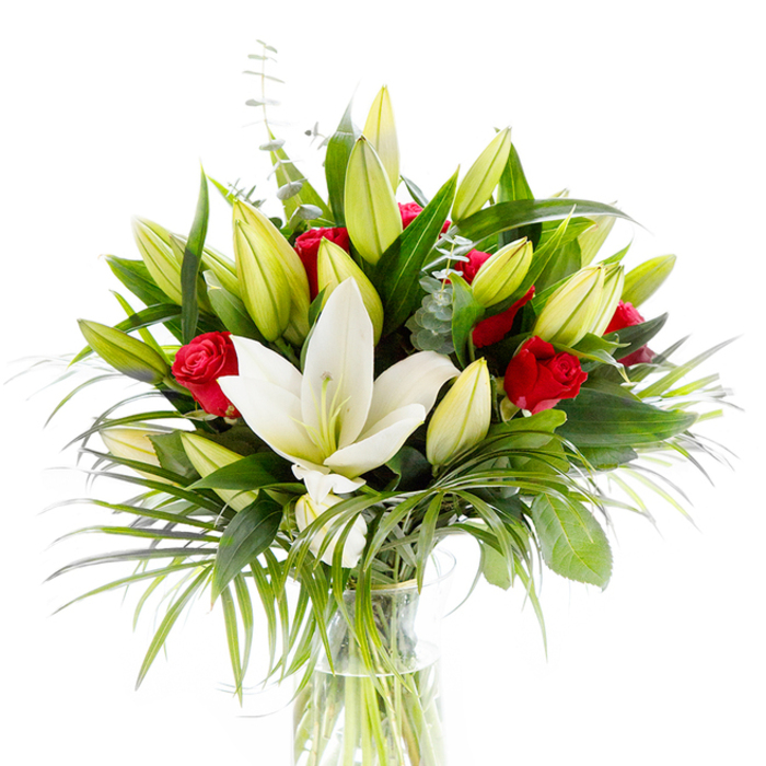 A striking, carefully balanced bouquet with a single, open creamy white Oriental lily placed slightly forward at its centre, its pristine petals unfolding to reveal a pale yellow stamen and a subtle green pistil. Ringing this focal bloom are many lily buds-soft lime, pale yellow and green-held at different heights to suggest growth and anticipation. Interlaced among the lilies, deep red rosebuds and small roses, mostly furled, supply rich, passionate punctuation that draws the eye across the arrangement. The floral mass is framed by an abundant palette of greens: broad, dark, glossy leaves that anchor the composition; long, arching palm-like fronds in a lighter emerald that introduce a sense of motion; and slender stems of silvery eucalyptus whose rounded leaves soften the overall texture. The bouquet sits in a simple, cylindrical clear glass vase; through the glass the stems are visible submerged in sparkling clear water, reinforcing a sense of freshness and transparency. Against a pure white background the colours and textures are crisp and readable-the lily's luminous whiteness, the roses' deep crimson, and the layered greens all emphasizing contrast and harmony. Imagine the combined scent, the lilies' heady sweetness mingling with the roses' velvety warmth, creating an atmosphere that is both celebratory and tender. Conceived with a florist's eye honed in Canonbury and neighbouring Islington streets, this arrangement reads as a refined gift for anniversaries, birthdays, or quiet moments of sympathy, equally at home as a dining table centrepiece or a heartfelt present delivered to a nearby home on a clear morning.