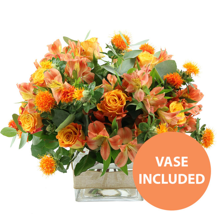 An opulent, autumn-leaning bouquet in rich oranges and yellows is shown against a bright white backdrop, every stem positioned with the care of an experienced Canonbury florist. Several large, deeply coloured orange roses form the visual anchor, their petals slightly variegated with hints of yellow toward the center, creating a feeling of depth and warmth. Interlaced with them are sunny yellow garden roses, their densely layered petals forming rounded, almost cushion-like blooms that radiate gentle cheer. Weaving through this base of roses are many stems of peach and light orange alstroemeria, or Peruvian lilies, the inner petals decorated with fine, striped markings that add a subtle pattern and sense of movement throughout the design. Standing out among the smoother textures are small, spiky, spherical orange accent flowers, reminiscent of safflower or ornamental thistles; their vivid colour and bristled form punctuate the arrangement with lively bursts of texture. Supporting the flowers is an abundant collar of foliage: soft, silvery-green eucalyptus leaves mixed with darker green fillers, giving the bouquet structure and the kind of full, generous silhouette you might place on a dining table in a townhouse off Canonbury Place. The stems are gathered neatly in a clear, square glass vase, its transparent sides revealing the clean lines of the stems and the clarity of the water, suggesting freshness and professional conditioning. Around the midsection of the vase, a wide band of natural beige burlap or jute is wrapped smoothly, lending a rustic, handcrafted feel that suits both modern apartments and period homes near New River Walk. At the bottom right of the image, a bold circular orange graphic with uppercase white lettering states VASE INCLUDED, clearly indicating that this is a complete, ready-to-display floral gift, ideal as a centerpiece for seasonal gatherings, thoughtful congratulations, or a comforting burst of colour on a sideboard overlooking the quiet streets of Canonbury.