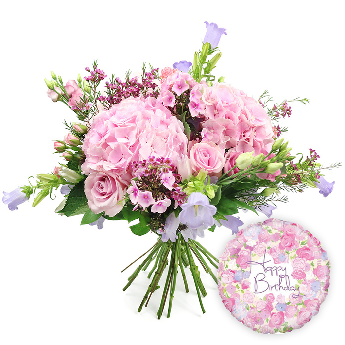 Set against a bright white background, a hand-tied birthday bouquet overflows with soft pink and lilac tones, giving the impression of a carefully arranged display ready to leave a Canonbury studio for same-day delivery. At the centre are several voluminous heads of blush pink hydrangea, each one made up of hundreds of tiny petals that together create plump, rounded shapes, like puffs of pastel clouds. Threaded among them are elegant soft pink roses, some fully open with layered petals curling back gracefully, others still in bud with tighter, more compact forms, adding structure and gentle movement throughout the bouquet. Smaller accents of deeper pink or fuchsia waxflowers, or similar miniature blossoms, are scattered between the larger blooms, their fine stems and clusters of tiny flowers offering contrast in both scale and colour. Rising here and there, slender stems of campanula display their bell-shaped flowers in light lavender and periwinkle blue, tilting upwards to introduce vertical interest and cool, airy tones that balance the warmth of the pinks. The entire composition is framed with abundant fresh foliage: broad leaves cradle the hydrangeas, while narrow, elongated leaves and delicate green stems soften the outline, helping the bouquet feel relaxed yet expertly composed. The stems beneath are clearly visible, neatly aligned and tied in the manner of a true hand-tied design. To the lower right of the flowers, a round Happy Birthday foil balloon rests, its reflective surface adorned with a repeating pattern of small roses and buds in shades of pink, cream, and lavender. Across its centre, the words Happy Birthday appear in a flowing purple script, echoing the romantic feel of the bouquet and suggesting a complete celebratory gift ideal for surprising someone in Canonbury, whether at home near Cross Street or in a local office.