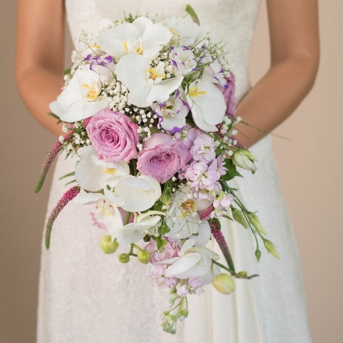 This expertly composed bridal bouquet, photographed against a muted background reminiscent of Canonbury's elegant Georgian terraces, draws the eye with its graceful waterfall shape and luxe harmony of colour. At the bouquet's heart, majestic white Phalaenopsis orchids unfold, each petal luminous and tender to the touch, clustered amid full-bloomed blush pink and lavender roses. Wisps of delicate Gypsophila soften the overall silhouette, adding a light, almost floating texture, while accents of slender Amaranthus in vivid magenta tumble dramatically downward. Lavender stock and sweet pea interlace gentle notes of colour, complementing the bouquet's flowing movement and natural taper. Sprays of green foliage and tight, unopened buds speak to the freshness and care with which Canonbury Flowers approaches bridal design. There is a sense of gentle perfume in the air-subtle, layered, and reminiscent of spring mornings along Canonbury Road. The bride's gown, with its fine lace detail and smooth, flowing skirt, completes the picture of refinement and joy. This bouquet is a visual and sensory celebration, ideal for those exchanging vows amidst Canonbury's leafy enclaves, embodying the artistry and devotion of local floral craftsmanship.