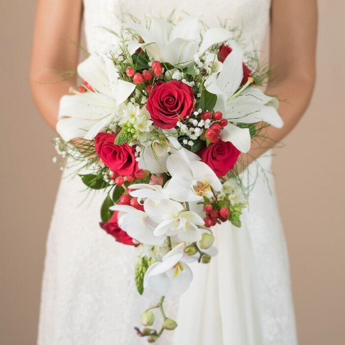 Against a serene backdrop reminiscent of Canonbury's peaceful avenues, a bride stands waist-up, her posture elegant as she carefully holds a lavish, cascading bridal bouquet in both hands. The floral artistry is striking-sumptuous white oriental lilies, petals pure and expansive, glow softly beneath diffused morning light, invoking a sense of calm and hope typical of a wedding day in Islington. Deep red roses, nestled between the lilies, reveal their tightly layered, velvety petals, their colour rich as autumn leaves in Duncan Terrace Gardens. Delicate white phalaenopsis orchids drape gracefully down the bouquet's length, each small cluster forming an intricate waterfall effect that mirrors the gentle flow of long-standing New River Walk. Sprigs of baby's breath and petite filler flowers shimmer between the main blooms, lending airiness, while small clusters of hypericum berries-bright and festive-dot the arrangement with bursts of vibrant red. Feathery green fronds and glossy broad leaves form a lush, textured base, punctuated by occasional spiky green accents that lend energy and modern structure. The bride's dress offers glimpses of fine lacework and smooth, satiny fabric, a reflection of traditional yet refined Canonbury wedding style. Every floral and textural choice harmonizes, resulting in a bouquet that is visually stunning and evocative of local charm-a timeless choice for couples seeking sophistication, romance, and a bespoke touch for their Canonbury ceremony.