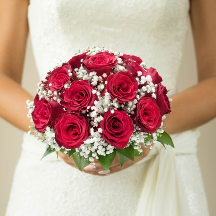 In a serene pocket of Canonbury, just steps from the tranquil New River Walk, a beautiful bridal bouquet takes centre stage in this close-up. Sensitively held at chest height, the bouquet is a testament to both tradition and local craft-a domed cluster of opulent, full-bodied red roses, their petals tightly layered and luminous, symbolising deep devotion. Draped among these passionate blooms, tiny white clouds of gypsophila weave softness and airiness throughout the arrangement, contrasting the roses' intensity with gentle purity. Tucked at the base, bright green leaves draw the eye, offering freshness and balance that echoes Canonbury's lush neighbourhood greenery. The bride's gentle, manicured hands embrace the stems, her fingers forming a protective cradle. Partially revealed above and behind, her gown shimmers in daylight: intricate lacework and a flowing sash create layers of visual and tactile delicacy, reflecting the neighbourhood's elegant history. The background, intentionally soft and neutral, ensures every detail of the artistry shines-perfect for couples in Canonbury seeking wedding inspiration, insight into local bridal trends, or a tangible glimpse into the heart of a meaningful celebration.