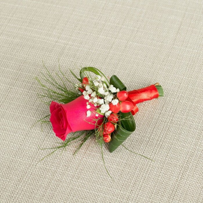 A meticulously arranged boutonniere rests on a gently textured, light beige background, the fabric resembling the kind of natural linen often seen in Canonbury homes and studios. At the centre, a vibrant fuchsia rose commands attention, its petals tight and velvety at the core, gradually unfurling in soft, rounded layers that suggest both freshness and emotional warmth. Encircling this central bloom is a gentle ring of white baby's breath, the tiny blossoms so numerous and delicate that they form a soft, cloud-like frame, brightening the overall palette and lending an air of quiet romance. Punctuating this whiteness are clusters of glossy red berries, likely hypericum, their smooth surfaces catching the light like tiny beads and echoing the passionate tones of the rose. Wispy sprigs of fine, feathery greenery extend beyond the main form, while broader dark green leaves are skillfully curled and layered to support the composition, adding structure and a sense of movement, as if the foliage is wrapping protectively around the rose. The stems beneath are hidden and held firmly by a wrap of rich red satin ribbon, its sheen contrasting beautifully with the matte texture of the linen and the natural surfaces of petal and leaf. The ribbon is finished in a neat, compact knot that feels both secure and refined, suggesting the professional hand of a florist well accustomed to dressing grooms and guests for Canonbury weddings and registry ceremonies. Soft, even daylight reveals every subtle shadow, highlighting the craftsmanship and making this small floral piece feel like a cherished token of joy ready to be pinned to a jacket lapel.