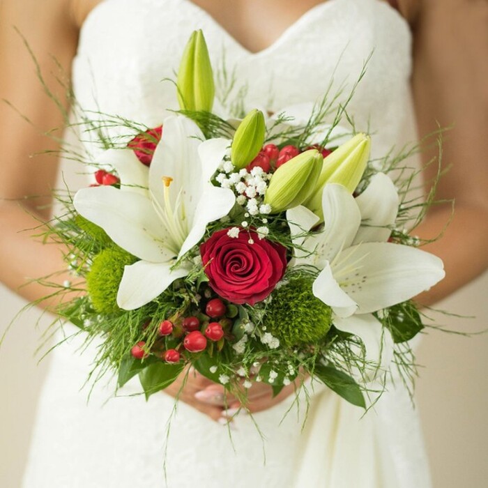 A close-up frame reveals a bride, poised with serene joy as she clasps a vibrant bouquet before her, all set against the gentle sheen of a white, subtly patterned wedding gown. The bouquet draws inspiration from Canonbury's harmonious blend of old-world charm and modern elegance, featuring open, snow-white lilies lit softly, their broad petals curving to reveal golden centres and casting gentle shadows that dance like the canalside leaves nearby. Amid these, velvety red roses capture the heart, their tightly furled buds evoking lasting romance, while bright green lily buds intersperse to signify fresh hope. Airy clusters of baby's breath create a light, floating texture, reminiscent of springtime petals caught on a breeze through Arlington Square. Red hypericum berries provide jovial splashes of colour, balanced by the cool, mossy touch of green trick dianthus or button chrysanthemums lending playful visual depth. Feathery asparagus fern and broadleaf greens peeking out suggest a bouquet freshly gathered from the community's parks and gardens. The composition is traditional yet lively, echoing Canonbury's unique blend of tranquillity and vibrancy. The image, softly illuminated and thoughtfully composed, would be a fitting icon for classic wedding ceremonies in local venues or for couples dreaming of floral designs rooted in their favourite North London neighbourhood.
