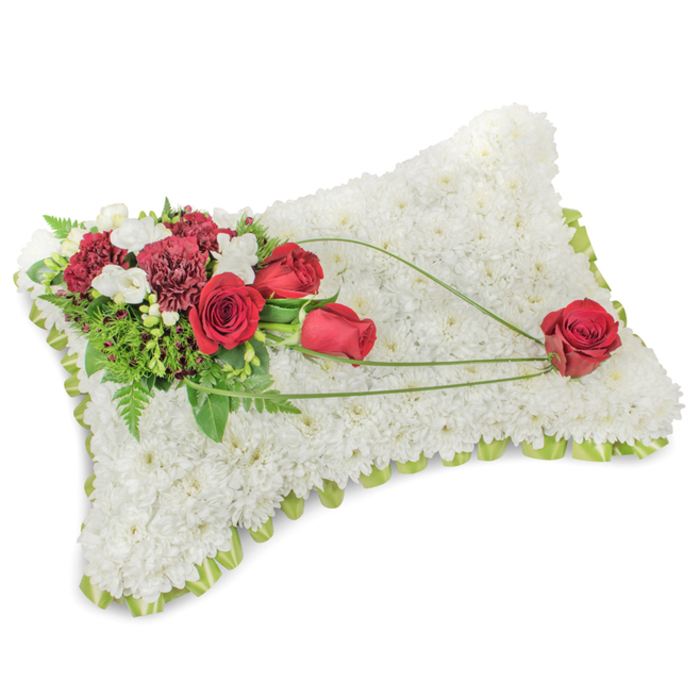 A classic rectangular funeral pillow composed entirely of dense white chrysanthemum blooms presents a restful, cloud-soft surface that evokes purity and consolation. The minute chrysanthemum heads are closely set to form a smooth, cushion-like plane, while a slim band of pale green ribbon or edging peeks out around the rim, offering a subtle, fresh definition to the silhouette. At the upper left, a carefully arranged floral spray combines deep red roses-some in bud, some unfolding-with rich burgundy carnations; their textures contrast beautifully, the roses' velvet petals against the carnations' frilled layers. Accents of tiny white freesias or spray chrysanthemums lighten the cluster and add a faint, sweet perfume. Supporting foliage of fern and glossy leaves provides shape and depth, giving the spray a living backdrop. From this corner, three slender lines of bright green bear-grass or decorative foliage sweep diagonally across the white field to the lower right, creating a graceful movement that culminates in a single, prominent red rose resting on the chrysanthemum surface. The overall impression is one of refined composition and gentle emotion-an offering of comfort and respect designed for services within Canonbury, from Canonbury Square to Essex Road, and for placement at local chapels, cemeteries or family tributes. The arrangement's balance of white serenity, vivid red sentiment and soft green accents reads as a timeless expression of remembrance and love.