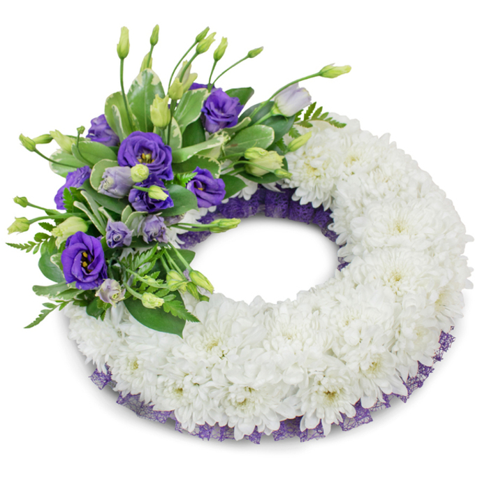 A serene, circular memorial wreath displayed against a crisp white backdrop, described here by a Canonbury florist who knows the quiet moments that follow a loss. The wreath's broad, continuous ring is assembled from tightly packed white cushion chrysanthemums, their ruffled petals creating a soft, even surface that reads as purity and repose. On the upper-left quadrant a concentrated spray of purple Eustoma, from deep violet to soft lavender, gives the composition an elegant focal point; some blooms are fully open with layered petals, others are delicate buds that suggest tenderness and unfolding memory. Variegated and glossy green foliage-intermingled with fern-like fronds-threads through the purple cluster, providing texture, natural contrast, and a sense of restorative greenery. Beneath the chrysanthemums a slim border of crinkled dark purple lace or mesh peeks out, tracing the inner and outer curves and echoing the lisianthus tones to complete the palette with a quietly regal flourish. The plain studio background and the soft shadow beneath the wreath emphasize its sculptural form and allow every petal and leaf to read clearly. There is a gentle scent and soft textural play in this arrangement-the crisp chrysanthemum cushion against the velvety lisianthus-crafted to express condolences. Prepared with local care by Canonbury Flowers for services in Canonbury Square, Essex Road chapels, or private remembrances, it stands as a lasting symbol of respect and love.