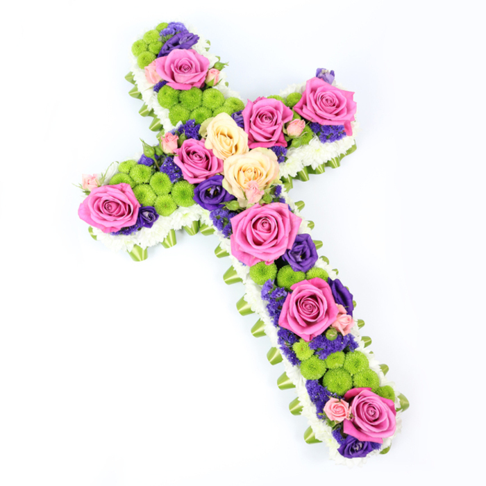 Set against a flawless white background, this floral cross appears as a luminous, carefully crafted tribute, gently tilted from upper left to lower right, giving the impression of being lovingly placed and thoughtfully photographed. The cross's outline is impeccably defined by a border of dazzling white spray chrysanthemums, each tiny flower head clustered tightly to form a smooth, cushioned edge, reminiscent of soft clouds drifting above Canonbury's rooftops. Just beneath this layer, a narrow band of folded light green ribbon, or perhaps neatly wrapped foliage, forms a second, discreet frame that subtly lifts the design and introduces a fresh, verdant tone. Within this border, the body of the cross is a vivid tapestry of colour and texture. Large roses in bold fuchsia pink mingle with softer tones of creamy pale yellow and delicate peach, their petals fully open and velvety, suggesting both the fullness of a life well lived and the tenderness of farewell. Bright, chartreuse button chrysanthemums sit in rounded clusters between the roses, their tight, spherical blooms adding a playful contrast to the larger, more romantic petals. Woven throughout, smaller flowers in deep purple and rich violet, likely lisianthus or statice, nestle into every gap, giving shadow and depth, rather like the layered planting in the gardens off Canonbury Square. The lighting is bright but gentle, illuminating each texture-the smooth satin of the ribbon, the crispness of the chrysanthemum petals, the softness of the rose blooms-creating a harmonious, balanced composition. This cross feels ideal as a Christian funeral tribute or memorial arrangement for a Canonbury service, conveying respect, remembrance, and a quiet sense of hope for those gathering to say goodbye.