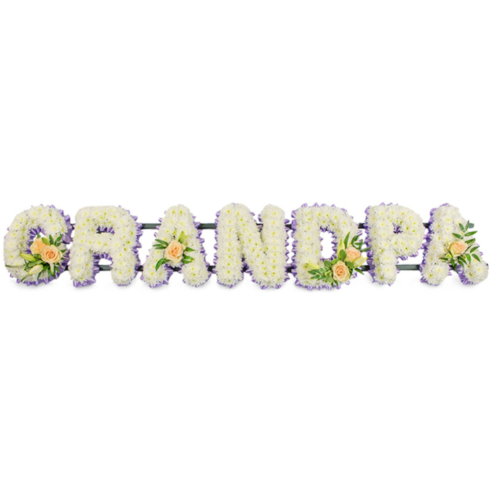 A long, freestanding floral arrangement spells GRANDPA in bold block capitals, arranged in a straight line as if resting on a low stand in a peaceful Canonbury chapel or local funeral home. Each letter is built from an abundant bed of white spray chrysanthemums, their small, closely packed blooms giving the surface a plush, cushion-like texture that appears soft and comforting. Around the outline of every letter runs a slender fringe of pale lavender and soft purple blossoms, which gently frame the white interior and bring a delicate contrast that makes the shapes easy to read from a distance. The letter G is enhanced with a carefully composed cluster of peach-toned roses, some open and some just unfurling, mingled with slim white lily buds and fresh green foliage that arcs naturally across the corner of the letter. Similar, but more modest, accents repeat through the word: a single peach rose and greenery on the first A, a slightly larger cluster on the D, and another single rose on the final A, creating a quiet rhythm of colour and life along the tribute. A dark horizontal support, likely a metal or painted frame, runs behind the base of the letters, connecting them while remaining visually discreet against the pure white background of the image. The overall mood is one of calm dignity and heartfelt affection, a bespoke funeral tribute that families in Canonbury might choose to lay beside the coffin or at a graveside service, expressing gratitude and enduring love for a grandfather whose presence is deeply missed.