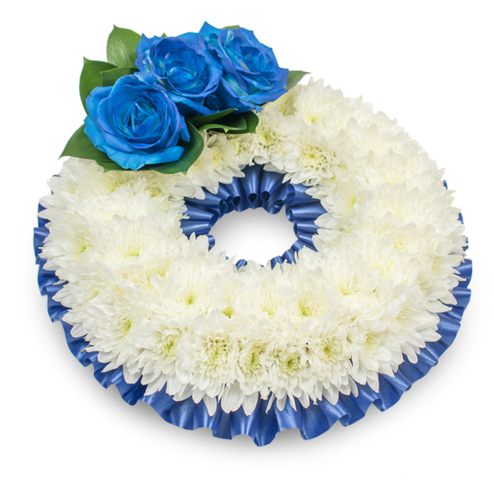 Captured against a bright white background from a modestly raised angle, this traditional circular wreath reads as an emblem of peaceful remembrance. The dominant texture is a thick, uninterrupted band of white chrysanthemums; their countless, feathery petals overlap to form a softly domed surface, and subtle creamy yellow hints at a few centres add warmth without disturbing the calm. Tucked at the upper left are three striking deep blue roses - their petals velvety and precisely unfurled into tight spirals, the intense sapphire tone offering a quiet, dignified contrast to the white. Dark green leaves nestle the roses and provide depth and natural framing. A pleated satin ribbon in matching rich cobalt blue borders both the inner and outer edges, its glossy folds catching light and shadow to give definition and an elegantly finished silhouette. The wreath casts a delicate shadow on the white plane beneath, which helps the arrangement read as three-dimensional and ready for placement. The scent is muted: a soft, comforting chrysanthemum fragrance that feels unobtrusive in a ceremonial setting. As a florist familiar with Canonbury cornerstones - from Chapel Market to St. Paul's Road - I would recommend this palette for funeral services or memorial gatherings where restraint and respect are required. The combination of classic white and profound blue communicates purity, solemnity, and a personalised note of tribute, suitable for laying during a service or leaving as a heartfelt sign of condolence.
