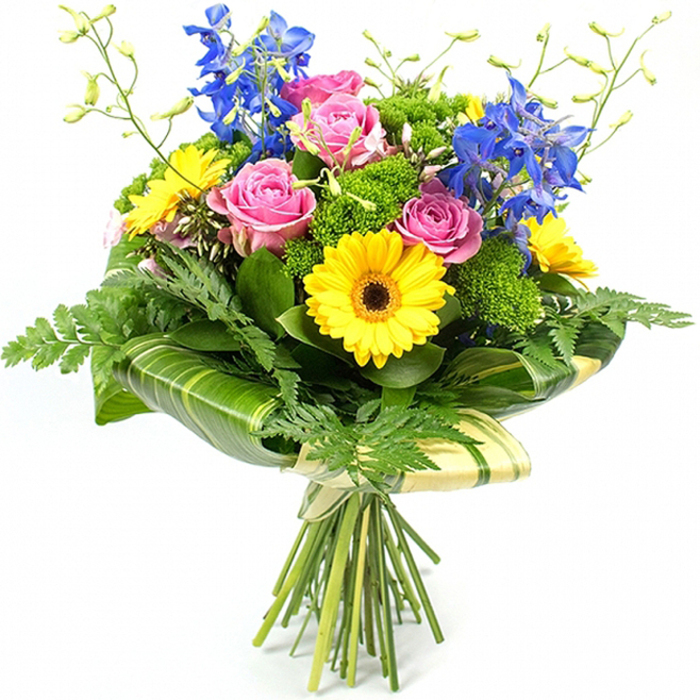 A richly detailed hand-tied bouquet is shown standing upright against a bright white backdrop, the simplicity of the background allowing every petal and leaf to take centre stage. Dominating the middle of the arrangement, several full, open pink roses reveal their layered, velvety petals, suggesting tenderness and affection with a classic, almost vintage charm. Around and between them, golden yellow gerbera daisies provide a sunny counterpoint, each bloom with a distinct dark chocolate centre that adds depth and visual focus, much like the lively pops of colour seen in Canonbury gardens in early summer. Towering above this warm heart of pinks and yellows, spires of deep blue delphinium rise with elegant poise, their multiple florets arranged along slim stems that add height and a cool, refreshing contrast. The bouquet feels abundantly green, with a variety of foliage types playing different roles: fine, feathery fern fronds soften the perimeter; dense, bright green filler foliage creates a mossy texture between blossoms; and broad, glossy tropical leaves provide structure and shine. Some of these larger leaves are skillfully curved and wrapped around the lower part of the design, creating a polished green collar that holds and frames the heads of the flowers. Slender green stems with tiny buds reach lightly upward, introducing an airy quality that keeps the arrangement from feeling heavy. All the stems are neatly gathered and tied with a natural-looking ribbon or twine, leaving the cut ends visible in a straightforward, hand-crafted style. The bouquet evokes the feeling of fresh, same-day flowers just delivered to a Canonbury townhouse, ideal for marking milestones, welcoming a new neighbour, or simply brightening a quiet corner near Essex Road.