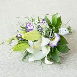 An elegant, elongated bridal-style bouquet of white lilies and purple lisianthus lies on a pale beige, subtly woven fabric, the neutral background allowing every detail of the flowers to stand out with clarity. The bouquet stretches on a gentle diagonal, fuller and more dramatic on the left, tapering to the neatly gathered stems on the right. Large Oriental lilies dominate the centre, their satin-like white petals opening wide to reveal warm yellow-orange stamens dusted in pollen and faint cinnamon-toned speckles scattered across the inner petals. Around these fully open blooms, smooth lime-green buds are tucked in, their pointed tips suggesting blossoms that will open in the coming days. Interlaced throughout are clusters of lisianthus, their ruffled petals tipped in rich purple that fades into creamy white centres, some blooms more tightly cupped, others open and airy, echoing the soft twilight tones often seen over Canonbury's tree-lined streets. Sprays of baby's breath add an ethereal finishing touch, with tiny white blossoms hovering lightly between the larger flowers. The greenery is lush yet refined: broad, dark green leaves with a gentle sheen provide a solid backdrop, while slender, wispy foliage and a few spikier stems introduce texture and a feeling of gentle movement. At the right-hand end, the long stems are bound in a smooth white ribbon, delicately accented with small pearl-like beads that hint at special occasions and quiet luxury. The soft, even lighting casts minimal shadows, giving the entire arrangement a tranquil, romantic atmosphere, ideal for a Canonbury wedding party, anniversary surprise, or elegant table display created by a local florist who understands the area's understated charm.