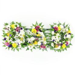 A beautifully composed funeral tribute spells out "DAD" in three bold floral letters, laid out in a neat row on a spotless white background so every colour reads clearly. Each individual letter is framed with a thick collar of glossy green fern, the feathery leaves giving a soft, garden-like feel and providing a strong outline for the shapes of the D, A, and D. Inside this leafy border, the letters are tightly filled with white chrysanthemums and wide-faced white gerbera daisies, creating a cushion of velvety white petals that suggests peace, purity, and quiet farewell. Nestled among the whites, clusters of yellow gerberas and petite yellow spray roses light up the tribute with a hopeful brightness, like the warmth of shared memories. In between, accents of purple lisianthus, deep pink or fuchsia spray roses, and small lavender-toned flowers weave through the design, offering subtle drama and conveying enduring love and respect. Sprigs of bright green button chrysanthemums and other soft green touches sit between the blooms, adding freshness and depth so the letters feel full and three-dimensional. Slim, dark support rails link the three designs, but they recede into the background, keeping the visual focus on the flowers and the heartfelt word they form. This arrangement feels perfectly suited to a Canonbury farewell, perhaps resting beside family tributes after a service near Highbury Fields or along a quiet church aisle, expressing gratitude and remembrance for a cherished dad in a way that words alone often cannot.