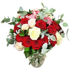 Set against a bright white background, this opulent rose arrangement feels like a captured moment of celebration, the sort of bouquet you might see arriving at a doorstep near Canonbury Square on an anniversary evening. The flowers spill generously from a clear glass vase whose rounded base and narrower neck cradle the stems, which are visible through the water and lend a natural, unforced structure. Deep red and scarlet roses dominate the composition, their petals plush and velvety, some blossoms fully unfurled in confident, romantic spirals, others still in bud, hinting at love and emotion continuing to open over time. Woven between these bold blooms are white and creamy ivory roses, along with subtle white roses kissed with blush pink at the tips, bringing light and softness into the arrangement so the reds seem even more intense. Here and there, small bright pink filler flowers cluster like confetti, their finer texture and petite size adding movement and a slightly playful note to the otherwise classic palette. Around and beneath the roses, blue-green eucalyptus stems with distinct rounded leaves fan outward, mingling with darker green foliage and the occasional silvery leaf to give the bouquet volume, depth, and a modern botanical feel. The eucalyptus suggests a cool, aromatic scent, the kind you'd notice when this design is delivered fresh from a Canonbury florist for same-day surprises in nearby Islington streets. The arrangement is rounded and full, expertly balanced so no single flower overwhelms the others, creating a luxurious yet approachable centrepiece, perfect for romantic milestones, elegant dinner parties, or heartfelt thank-yous.