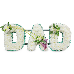 The image presents a lovingly crafted funeral tribute spelling "DAD" in sturdy three-dimensional letters, a design that would feel at home at a quiet Canonbury service or a farewell near Highbury Corner. Each letter is completely carpeted in crisp white chrysanthemums, their tight, button-like blooms placed so closely that no gaps appear, creating a plush, almost velvety texture. They catch the light softly, giving the tribute a calm, luminous quality against the plain white background. A rich emerald green satin ribbon, carefully pleated and ruffled, outlines every curve and corner of the letters, bringing definition and a touch of natural colour reminiscent of the tree-lined streets around Canonbury Square. Behind the flowers, the dark horizontal lines of the support frame peep through just enough to show how the structure stands upright as one united piece.   

Thoughtful accent flowers break the white expanse in just the right places. At the upper left of the first "D", a small spray of white freesias and glossy green leaves emerges, their slender buds and open florets creating a sense of gentle movement. Two elegant green stems sweep diagonally from this first "D" across towards the centre of the "A", guiding the eye to a striking focal arrangement. There, two deep purple calla lilies nestle among more white freesias and foliage, their rich, sombre hue adding depth and a dignified contrast. On the final "D", a more compact cluster of freesia and greenery mirrors the first, completing the visual balance.   

This "DAD" tribute conveys quiet strength, gratitude, and sorrow, perfectly suited for a father's funeral, wake, or memorial gathering around Canonbury, whether placed beside a coffin, at a graveside, or near a display of family photographs.