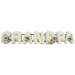 A lovingly designed funeral tribute spells out GRANDPA in bold, three-dimensional floral letters, laid out in a straight line against a clean white backdrop that mirrors the soft light of a peaceful Canonbury morning. Each capital letter is completely filled with tight clusters of white chrysanthemums or similar small-petalled flowers, so densely packed that the surface looks almost like freshly fallen snow, smooth yet gently tufted. Around the edges, a fine border of lilac and light purple blossoms picks out the shape of each letter, their tiny star-like petals giving a subtle, feathery fringe that adds definition without overpowering the serenity of the white. Several letters hold small, meaningful posies: the 'G' carries a graceful white lily-like flower rising from a nest of fuchsia and blush-pink blooms; the first 'A' and the last 'A' each display petite clusters of purple flowers, set against dark, polished greenery; the 'D' features a more prominent spray that combines pink flowers, another elegant white lily form, and bright green button chrysanthemums that lend a touch of freshness. A slim, dark green frame runs quietly behind the letters, keeping them level and secure, as if ready to rest near a family pew in a church off Essex Road or be carried to a memorial gathering close to Canonbury Gardens. The smooth white background removes any distraction, drawing the eye to the textures of petal and foliage, the gentle contrast of white and lavender, and the careful balance of colour. The tribute conveys a sense of warmth, gratitude, and respect, an intimate way to say goodbye to a cherished grandfather while surrounding his memory with softness and light.