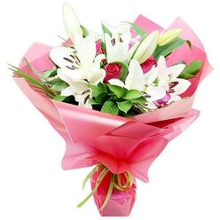 A sophisticated bouquet of white lilies and deep red roses is shown against a simple white background, capturing the careful craftsmanship of a local Canonbury florist. At first glance, your eye is drawn to the impressive Oriental lilies, some fully open with wide, trumpet-like petals and others still elegantly closed, their buds a pale green brushed with ivory. The open blooms reveal central clusters of dark, almost purple-brown anthers on slender filaments, adding rich contrast to the luminous white petals. Threaded throughout the lilies are velvety red rosebuds, some tightly coiled and others beginning to reveal their layered hearts, evoking romance and devotion. Around this floral duet, an abundance of deep green foliage creates a lush frame: broad, polished leaves catch the light with a gentle sheen, while thin, arching blades of grass-like greenery give the arrangement a soft, flowing silhouette. The entire bouquet is wrapped in a voluminous layer of glossy bright pink cellophane or floral paper, which billows and folds like fabric, turning the hand-tied stems into a beautiful, ready-to-give gift. At the base, a delicate lime green ribbon is tied in a small knot, its fresh tone echoing the natural greens and lending a modern twist. The design feels both elegant and joyful, the kind of bouquet that might arrive as a same-day delivery to a home near Canonbury Station, a cosy restaurant off Upper Street, or an office close to New River Walk, bringing with it not only fragrance and colour but a clear message of appreciation, love, or celebration.