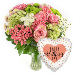 A bright, celebratory Mother's Day gift arrangement is shown in a clean studio setting, the pure white backdrop echoing the light townhouses and leafy calm of Canonbury's residential streets. At the centre of the image stands a rounded bouquet in a clear glass vase, brimming with fresh blooms that lean toward soft springtime tones. Several large pink roses form the heart of the design, their petals opening in generous spirals, tinted with deeper pink toward the centre and softening to delicate, almost cloud-like edges. Around them, clusters of coral-pink blossoms, similar to kalanchoe, create a lively, textural ring, each tiny flower catching the eye with its compact, starry shape. Lime green button chrysanthemums nestle among the pinks, their spherical, almost tufted heads adding a modern, slightly quirky contrast that feels both fun and elegant. Sprays of small white, daisy-like chrysanthemums weave through the composition, bringing lightness and balance, while rich green foliage fills out the base, giving a sense of natural abundance. The stems, visible through the vase, are neatly cut and immersed in clear water, reinforcing the impression of freshness you'd expect from a same-day Canonbury delivery. Just to the lower right of the bouquet floats a lustrous heart-shaped foil balloon. Its white or pale silver background is dotted with irregular polka dots in gold, silver, and soft peach, like confetti drifting through the air. At its centre sits a solid peach-coral heart carrying the phrase HAPPY Mother's DAY in elegant black lettering. Together, the flowers and balloon convey warmth, appreciation, and happy anticipation, as if ready to be carried up a narrow Canonbury terrace staircase and placed on a kitchen table for a well-loved mum.