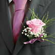 This image offers a detailed close-up of a man's upper torso dressed for a special event in Canonbury, perhaps a wedding at a local church or a formal celebration near the tree-lined streets off St Paul's Road. His tailored suit is a deep charcoal grey, almost black, with a smooth, structured lapel that frames the focal point of the photograph: a thoughtful, handcrafted boutonniere. Underneath the jacket, a light dress shirt patterned with a fine check adds a subtle, almost textured backdrop, while a striking fuchsia or magenta tie runs neatly down the centre. The tie's silky finish reflects the available light, infusing the scene with a rich, berry-tone warmth that feels both modern and romantic. Pinned on the left lapel, the boutonniere features a single pale pink rose, its petals soft and velvety, opening in gentle spirals from a deeper rosy centre to paler, almost blush edges. Surrounding the rose, delicate stems of white baby's breath form tiny clusters of blossoms, like small puffs of cloud or lace trim, bringing a bright, airy lift to the composition. Wispy green foliage rises behind the main flower, slim and lively, suggesting fresh, just-cut stems from a local florist. At the base of the arrangement, a broader green leaf or wrapping gathers the stems, finished with a satin bow in muted purple or lavender that echoes the tie's pink notes without overwhelming them. The entire ensemble conveys affectionate celebration and careful preparation, as if this is the final touch added just before stepping out into a day filled with toasts, photographs, and cherished memories along the quiet streets of Canonbury.