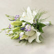 This fresh, ethereal bouquet rests horizontally on soft beige linen, its arrangement composed with a studied sense of restraint and grace. Dominating the scene are immaculate white lilies: several trumpet-shaped blooms fully open, their silky petals curving back to expose luminous creamy centers, golden stamens, and the occasional delicate speckling within each bloom; nearby, poised green buds suggest unfolding promise. Interspersed among them are white lisianthus whose ruffled petals bear a subtle lavender-purple halo along the edges, offering a gentle whisper of colour that lifts the predominantly white palette. The texture is animated by slender, fern-like greenery that threads through the flowers, adding a featherlight movement as it catches the soft directional light and casts faint, natural shadows. Deeper green leaves anchor the bouquet, providing structure and contrast to the lighter elements, while small sprays of baby's breath are woven discreetly among the blooms, contributing an airy, cloud-like softness. The stems converge to the right, bound in a smooth white satin ribbon tied with care - a finish that reads both modern and timeless. The composition, photographed in diffused daylight, feels like an early-summer morning in Canonbury near Essex Road and the quiet edges of Regent's Canal: restorative, clean, and quietly celebratory. It would be equally appropriate as a bridal bouquet for an intimate chapel ceremony, a refined gift to mark an anniversary, or as a tasteful arrangement sent to convey sympathy, each use guided by the same sense of measured elegance and floral craft.