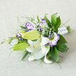 On a light, oatmeal-toned fabric that looks like softly woven linen, a bridal bouquet lies gently on its side, as if just set down between photographs on a Canonbury townhouse staircase. The composition is led by white Oriental lilies, their long petals arching back in a graceful curve, with some blooms completely open and others still in sculpted green buds. Inside the open lilies, warm yellow stamens tipped with burnished brown pollen add depth, while delicate burgundy freckles decorate the inner petals like fine lace. Woven among these statement flowers are lisianthus in white and rich violet, each bloom showing a creamy centre that gradually deepens to purple along the ruffled edges, creating a painterly, almost theatrical effect. Smaller lisianthus buds, still closed with a hint of lilac, bring texture and suggest that the bouquet will keep evolving throughout the celebration. Airy clusters of pure white baby's breath form a gentle halo, softening the lines of the larger blooms and giving the design a cloud-like lightness. Surrounding everything is an abundance of greenery: broad, polished leaves that catch the light, finer feathery foliage that hints at woodland ferns, and slender grasses that extend slightly beyond the blooms, adding a loose, garden-inspired silhouette. At the base, the stems are carefully aligned and wrapped in white satin, the ribbon smoothed tight and dotted with tiny, gem-like pearls that glint discreetly, echoing bridal jewellery. This refined yet natural bouquet, typical of the hand-crafted work created daily for Canonbury weddings, feels perfect for ceremonies in nearby Highbury or strolls to celebrations along Upper Street, conveying purity, devotion, and the hopeful excitement of a new chapter together.