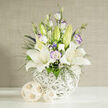 In this carefully lit image, a refined white and purple bouquet rests on a clean white surface, as though just set down in a calm Canonbury sitting room. Several majestic white lilies dominate the arrangement, their petals fully unfurled and silky, framing speckled throats and warm, pollen-rich stamens tipped in amber. Threaded between these lilies are graceful stems of lisianthus in pure white and soft lilac, some blooms open with frilled, almost paper-like petals edged in violet, others still curled tightly in pale green buds that lend a sense of gentle anticipation. Fine, dark green foliage arcs and sways through the bouquet, interlaced with dainty clusters of white filler flowers and tiny, jewel-toned blossoms in deep blue and purple, adding depth and a subtle, contemporary edge. The flowers are gathered into a distinctive round vase crafted from overlapping white strands, its woven, basket-like structure casting delicate shadows that highlight the sculptural shapes of each bloom. To one side, two small decorative spheres made of blonde, rattan-like material sit in quiet harmony, their natural threads echoing the vase's texture. Behind, a softly woven beige backdrop suggests the understated elegance of a Canonbury townhouse interior, calm and inviting. The composition feels balanced and poised, ideal for a refined wedding breakfast near Highbury & Islington, a peaceful condolence bouquet, or a stylish accent on a dining table overlooking New River Walk, all prepared with the practiced hands of a local florist who understands both design and emotion.