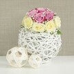 In this serene floral scene, a rounded bouquet of roses sits gracefully atop a white woven vase, evoking the atmosphere of an intimate Canonbury wedding or a carefully styled celebratory table. The arrangement is compact and symmetrical, made up of around a dozen fresh roses clustered closely together to form a smooth dome. Soft pink blooms with a hint of lavender nestle against creamy ivory and pale yellow roses, their colours blending like watercolour on paper. Some petals remain wrapped tightly around their centres, while others are gently open, suggesting flowers cut at their peak and arranged with practiced hands. Scattered throughout the roses are delicate sprigs of white baby's breath, their tiny star-like flowers creating a misty veil around the main blooms and adding a timeless bridal touch that would look at home in any Canonbury townhouse or nearby garden venue. The bouquet rests in a distinctive, spherical vase crafted from slender white wicker or painted twigs, intricately woven into an open, cage-like globe. The airy design lets glimpses of the inner container show through and introduces a sense of lightness and craftsmanship. This vase stands on a spotless white surface, perhaps a dining or reception table set for guests. To the left of the main arrangement, two decorative spheres made from pale, natural wood or rattan echo the woven theme in smaller, more minimal forms, one larger and one small, providing rhythm and balance. Behind the display, a textured beige or light taupe background, reminiscent of linen or fine wallpaper, casts a warm, neutral glow. The overall feeling is one of quiet romance and tasteful simplicity, perfect for illustrating local wedding table arrangements, anniversary celebrations, or refined home décor styling across Canonbury.