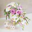 A romantic floral basket, perfect for a Canonbury wedding or engagement celebration, is shown resting on a polished white tabletop that subtly mirrors its shapes and colours. The base is a white wicker basket with an intricate, close-knit weave, lending a tactile, hand-crafted feel that suits the quiet, village-like character of the Canonbury streets. From this basket, flowers spill in a loose, asymmetrical cascade, creating the impression of a garden carefully gathered and artfully arranged. At the centre, large pink roses dominate, each bloom fully unfurled with thick, velvety petals spiralling towards a soft heart, giving the arrangement a rich, romantic core. Around them, white Phalaenopsis orchids arc outward, their broad, flat petals and delicate yellow centres adding an elegant, contemporary note. White lisianthus flowers, some brushed with a hint of lavender on the ruffled edges, mingle through the roses and orchids, deepening the sense of texture and fullness. Light, cloudlike sprays of white gypsophila fill the gaps, softening the transitions between blooms. Along the outer edges, smaller, soft pink flowers, reminiscent of stock or Sweet William, trail gently downward and emphasize the cascading form. A tall, narrow spike of deep purple-magenta veronica pierces the arrangement, adding graceful height and a touch of bolder colour. Fresh green foliage tucks throughout, providing structural support and a fresh, garden-inspired backdrop. In front of the basket rest two decorative, openwork spheres made from pale, natural rattan or wood fibre, one noticeably larger than the other, their lattice-like design echoing the basket's weave. A soft, textured taupe fabric drapes in the background, diffusing the light and keeping the mood calm and refined. The scene feels like a quiet moment in a Canonbury townhouse-elegant, heartfelt, and ready to be delivered locally as a centerpiece or a cherished gift.