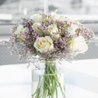 A tranquil sympathy bouquet of white roses and soft purple statice fills the frame of this image, composed with a tenderness that feels deeply appropriate for moments of remembrance. At the heart of the arrangement, several creamy white roses have opened fully, revealing intricate spiral centres and gently ruffled petals that seem almost satin to the touch. The colour is a restrained blend of white and ivory, suggesting purity, new beginnings, and a calm acceptance all at once. Woven between the roses are feathery stems of lavender, lilac, and pale pink statice, their many tiny blossoms forming a light, cloud-like surround that softens the stronger presence of the roses. The contrast of smooth, velvety petals against the more textured, papery filler flowers lends the bouquet an inviting depth. All of the stems are gathered in a tall, clear glass vase, the fresh green lines of each stem visible through the water, echoing the simple, uncluttered style often seen in Canonbury homes and studios along Upper Street. The vase rests on a clean white surface that catches and diffuses the natural light, as though the flowers were placed near a bright window before being carried out for delivery. In the softly blurred background, only gentle neutral tones can be made out, ensuring that nothing distracts from the quiet beauty of the blooms. The overall mood is calm, reflective, and sincere, making this arrangement an ideal way to express sympathy, offer support to grieving families in Canonbury, or bring a sense of peaceful presence to a memorial or condolence visit.