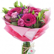 A joyful hand-tied bouquet sits upright against a clean, brilliant white backdrop, its energy focused in a compact, artful dome of colour. At the centre, several large gerbera daisies blaze in a rich fuchsia pink, each with a deep, almost black eye that sharpens the petals' vivid tones. Around them, pale pink roses appear in stages of opening - from tight, elegant buds to softly unfurling blooms whose layered petals create a gentle, concentric rhythm. Sprays of deep purple-blue statice (or similar filler) are threaded between the roses and gerberas, adding a fine, textured punctuation that draws the eye inward. Lush green foliage - broad glossy leaves and slender stems - forms a verdant cradle that gives the composition depth and a natural, garden-fresh vitality. The flowers are wrapped in layered, crinkled pink cellophane or tissue that graduates from an almost translucent pastel pink near the top, forming a soft halo, to a brighter, more saturated pink lower down; the glossy surfaces catch the studio light and lend a celebratory gleam. The bundled stems are visible through a clear cylindrical glass vase which holds the bouquet with casual confidence; a length of rustic twine is tied around the vase neck and from it dangles a small white rectangular tag printed with a stylised pink and green rose logo, suggesting a local florist's signature. The overall impression is fresh and uplifting - a perfect floral choice for birthdays, Mother's Day, anniversaries, or to brighten a Canonbury kitchen table on a spring morning, carrying the scent of a neighbourhood florist's shop into the home.