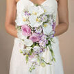 In this detailed waist-up wedding image, a bride stands quietly against a smooth, neutral beige background, her white sweetheart neckline dress catching the light in gentle highlights that frame the bouquet she holds. The arrangement is a luxurious cascading bridal bouquet that falls in a soft teardrop, rich with fresh flowers in white, shades of blush and rose pink, and muted lavender tones. Dominating the centre, wide petals of white Phalaenopsis orchids unfurl like silk, their yellow-orange centres forming tiny glowing hearts that draw the eye. Around them, full-headed pink roses and dusky lavender-pink garden roses create depth and romance, their petals layered in intricate spirals that speak of time and care. Sprinkled throughout are clusters of dainty white lisianthus, some petals brushed with the faintest hint of purple, and fine sprays of white gypsophila that seem to float like small clouds. Spires of light lavender stock weave between the larger blooms, adding a slightly wild, garden-like feel and hinting at a cool, sweet scent you might notice walking down Canonbury's tree-lined streets on an early spring day. The bouquet flows downwards in elegant stages, each flower placed to continue the line of the cascade without feeling stiff, giving the impression of movement even in a still photograph. Subtle green stems and leaves provide a quiet framework, almost hidden yet essential to the structure. This image beautifully captures a refined yet romantic bridal look, perfect for illustrating wedding flower inspiration, pastel colour palettes, or the type of graceful bouquets chosen for intimate ceremonies in Canonbury and neighbouring Islington.