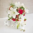 A sophisticated floral display, ideal as a wedding table arrangement or special celebration centerpiece, is arranged on a smooth white surface before a softly textured beige background that could easily belong to a Canonbury studio filled with natural light. The flowers are gathered in a rounded white basket-style container with an intricate open weave, giving a handcrafted, airy feel that contrasts gently with the clean tabletop. Emerging from this base, several generously sized white oriental lilies open in full splendour, their broad petals curving outward in star-like shapes and revealing prominent stamens that add a subtle golden note. Between and around the lilies, velvety red roses sit in rich, saturated tones, their tightly layered petals adding a sense of depth, devotion, and quiet drama. Wisps of white baby's breath thread throughout the bouquet, lending a floaty, cloud-like softness that balances the bolder blooms, while fine, bright green, fern-like foliage introduces movement and a fresh garden character. Scattered among these elements, small clusters of glossy red berries, likely hypericum, punctuate the arrangement with playful, festive accents that hint at winter celebrations or romantic evenings near Canonbury Square. From the right-hand side, a spill of white Phalaenopsis orchids extends gracefully outward and slightly downward, their arched stems carrying several open blooms with soft yellow and pink centres, along with a few poised buds that promise future blossoms. On the left side of the scene, three delicate white woven spheres rest on the tabletop, echoing the container's texture and giving the entire composition a sense of thoughtful balance, refined craftsmanship, and timeless elegance.