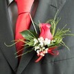 The photograph captures a single, finely crafted wedding boutonniere pinned neatly to the lapel of a dark charcoal grey suit, as if ready for a walk from Canonbury over to a celebration on Upper Street. At its heart is a rich, deep pink rosebud, half-open so that its velvety petals reveal a gentle spiral, glowing softly against the darker fabric. Encircling the rose, delicate white baby's breath forms a airy collar of tiny blossoms, giving a light, cloudlike contrast that speaks of quiet romance. Slender, feathery green sprigs and a few smooth, broader leaves fan out behind the flowers, adding a touch of natural movement and a fresh, garden-inspired feel that echoes the greenery of nearby Canonbury Square. The stems are wrapped and finished at the base with a bright red satin bow, its gleam mirroring the bold red silk necktie that lies neatly beneath the suit's lapel. The suit material itself looks like a fine wool blend, with a subtle, refined texture, while a lightly patterned grey shirt shows just enough to create depth without competing with the floral accent. Gentle, even lighting bathes the scene, highlighting the interplay of colour and texture-from the sheen of the ribbon to the matte softness of the rose petals-without any harsh glare. The overall impression is one of thoughtful coordination and understated luxury, a buttonhole designed for a groom or groomsman who appreciates detail and wants their wedding flowers to complement Canonbury's blend of tradition, style, and intimate city charm.