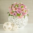 In a softly sunlit room characteristic of Canonbury's charming Victorian homes, a show-stopping table centerpiece radiates both freshness and creative flair. This handcrafted bouquet is anchored by a unique, white vase of intricate open-weave design-its slender, interlaced strips creating a captivating texture similar to artisan pieces seen along St. Paul's Road. The vase's shapely, rounded form is sturdy and inviting, cradling a profusion of pastel pink and lavender roses in full, velvety bloom. These classic flowers are thoughtfully accented with elegant pink alstroemeria, their trumpet-like petals patterned with gentle stripes and subtle touches of yellow. Clusters of playful, lime green button chrysanthemums-lively and compact-add vibrancy, while gossamer sprigs of baby's breath impart ethereal, airy softness across the bouquet. Dark green leaves peep through, grounding the lighter tones and offering richness. To the left, two decorative spheres made from pale woven material-one large, one smaller-reflect the latticework of the vase and lend the arrangement visual balance and a modern, textural appeal. Everything rests on a pure white tabletop, with a calm, textured light beige wall behind, allowing the bouquet's gentle colours and thoughtful craftsmanship to take centre stage. This scene captures the feeling of quiet celebration and creative expression, whether for elegant weddings, anniversary dinners, or simply brightening up homes throughout Canonbury with a touch of local floral artistry.