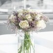 A rounded bouquet of creamy white roses and lilac statice stands in the centre of the image, arranged with the quiet confidence of an experienced Canonbury florist. Around a dozen full, velvety roses, the colour of warm ivory with just a hint of blush, open gently so that each layered petal is visible, forming soft spirals that draw the eye inward. Threaded between them are airy clouds of tiny lavender-purple statice blossoms, their papery texture and fine stems creating a mist-like halo that softens the edges of the bouquet. The flowers sit in a tall, cylindrical clear glass vase, its smooth surface catching the light and revealing the straight green rose stems submerged in clear water, some stems slightly crossing like lines in a gentle sketch. The vase rests on a bright white tabletop that reflects the arrangement in a faint, blurred glow, adding to the impression of freshness and cleanliness. Behind, the background is softly out of focus, washed with pale daylight as if coming through a large Canonbury townhouse window, suggesting an elegant indoor setting close to Islington's leafy streets. The light glances off the rose petals, making them appear almost luminous, while the lilac sprigs cast the faintest shadows. There are no other objects to distract from the bouquet; the entire scene feels composed for contemplation and calm. The overall mood is serene and uplifting, ideal for a sympathy gesture, a quiet anniversary in a North London flat, or a thoughtful delivery that brings a gentle breath of garden scent and refined beauty into a modern home.