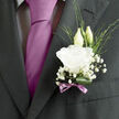 Pinned close to the heart of a dark wedding suit, this boutonnière looks like a delicate promise shaped by an experienced florist's hands. A single white lisianthus bloom unfurls gracefully at the centre, its petals soft and slightly frilled, layered in loose spirals that recall fine crepe fabric or the folds of a bridal gown. Behind it, two elegant buds stretch gently upward, pale green and ivory, poised between opening and stillness, capturing that sense of expectation that lingers just before vows are spoken. Surrounding the focal bloom, sprigs of baby's breath scatter tiny white blossoms like a fine mist, adding lightness and an almost cloud-like halo around the lisianthus. Slim strands of greenery weave through the design, bringing gentle movement and a natural, garden-picked feel that would be perfectly at home in a ceremony just off Upper Street or in one of Canonbury's hidden garden squares. At the base, a rolled, deep-green leaf curves in a neat loop, creating a clean frame that holds the composition together. The stems are bound with a narrow mauve ribbon, tied into a careful bow whose soft sheen echoes the groom's tie or pocket handkerchief without overpowering the flowers. The colours remain calm and understated-white, green, and a muted ribbon tone-yet the effect feels quietly confident, refined, and deeply personal. You can almost sense a gentle, powdery floral scent and the slight rustle of petals against fabric as the groom walks, carrying with him a subtle symbol of commitment and new beginnings.