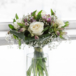 A soft and contemplative white and lavender bouquet stands in a clear glass vase, set before a luminous, gently blurred background that feels like a bright yet hushed Canonbury interior. At its heart, fully opened white roses display layers of plush petals, each bloom shaped into a perfect rosette with a creamy ivory centre that seems to glow. Surrounding them, clouds of white gypsophila drift between the roses, the countless tiny blossoms creating a weightless, mist-like texture that gives the whole arrangement a floating, ethereal quality. Woven through are stems of alstroemeria, their narrow buds and partially open flowers tipped in muted lavender and pale purple, some petals marked with fine, natural striping that adds quiet complexity rather than drama. Encircling the bouquet, broad, dark green leaves are expertly folded and curved, forming a polished collar that supports the rounded, hand-tied form and grounds the lighter tones above. The cylindrical glass vase is perfectly clear, revealing the straight green stems gathered together and resting in fresh water, the waterline catching a subtle glint of light. Everything about the composition feels measured and intentional, as though a Canonbury florist has arranged it with particular care for a condolence delivery along St Paul's Road or a reflective corner near Canonbury Station. The mood is one of gentle strength and understated grace, making this arrangement a fitting choice for funerals, sympathy messages, or quiet memorials where soft, elegant flowers can offer comfort, presence, and a sense of peaceful remembrance.