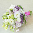On a pale, textured beige fabric backdrop, a hand-tied bouquet is positioned on a gentle diagonal, its colours unfolding in a sophisticated blend of white, lavender, purple and soft green, much like a carefully curated window display at a Canonbury florist. Lush white lisianthus blossoms form the focal point, their velvety petals layered in generous ruffles around creamy yellow centres, while nearby buds-elongated and tinged with pale green-hint at blooms still to come. Between these classic flowers, airy tufts of lavender-pink florets and clusters of star-shaped purple allium create a subtle contrast, their spiky, sculptural forms lending a modern twist to the traditionally romantic composition. The greenery is abundant yet restrained, with long, slender leaves, delicate loops of grass, and lighter green textural elements weaving rhythm and movement through the bouquet, echoing the natural, slightly wild feel of a walk through the leafy streets near Canonbury Station. The stems are meticulously aligned and bound with a bold fuchsia satin ribbon that wraps smoothly around the handle, further embellished with a fine strand of white pearls that catch the light with understated glamour. The result is a bouquet that feels both timeless and current, as suitable for a chic Canonbury townhouse wedding as for a special birthday or engagement celebration delivered across nearby Highbury and Islington. Although the image is silent, you can almost sense the gentle fragrance of the lisianthus and garden flowers, and feel the cool satin and smooth pearls against your palm, capturing a mood of new beginnings, thoughtful craftsmanship, and bespoke elegance from a local North London perspective.