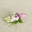 A delicately crafted floral boutonniere rests on a softly textured, oatmeal-toned linen background, the neutral fabric resembling the kind of elegant table linens you might see at an intimate Canonbury wedding venue. At the centre of the piece is a compact cluster of ruffled, soft pink blossoms, similar to miniature stock or dainty garden flowers, their petals layered and slightly frilled, with gentle gradients of blush and rose along the edges. Threaded between these pastel blooms are airy clouds of tiny white baby's breath, each floret crisp and bright, adding a lace-like lightness to the design. Nestled among the pink and white flowers are a few discreet accents of tiny lavender and deep purple florets, bringing a subtle depth and hint of romance, like the twilight sky over nearby Highbury Fields. On one side, several creamy white to pale green buds, suggestive of freesia or another slender bloom, extend gracefully outward, still closed and full of promise, evoking the feeling of a special day just about to begin. The flowers and buds are framed by lush, broad green leaves with a rich, emerald sheen, providing structure and contrast, while fine, wispy strands of grass-like greenery and ferny sprigs reach outward in soft arcs, giving a natural, slightly wild edge. At the base, the stems are neatly bound and wrapped in a vivid fuchsia satin ribbon, tied into a perfectly formed bow with smooth, looping tails that lie gently against the linen. The lighting is soft and even, highlighting the freshness and craftsmanship, making this boutonniere feel ideal for a Canonbury ceremony, a prom near Upper Street, or any refined celebration where a handcrafted floral detail speaks quietly of care, joy, and thoughtful design.