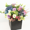A striking, modern creation from a Canonbury florist, this lushly abundant bouquet is presented in a sleek black square box that offsets its vibrant display of flowers. Lavish clusters of deep indigo and periwinkle blue hydrangeas dominate the rounded shape of the design, their fullness evoking the manicured gardens and green squares scattered throughout Canonbury. Interwoven among these are elegantly spiraled roses in shades of lavender and mauve, their delicate petals gently opening to invite closer admiration. Creamy white lisianthus blooms contribute an air of classic elegance, while bursts of fuchsia-pink filler flowers and subtle dark berries introduce playful color and fine texture. Varied greenery, from bright lime accents to glossy, darker leaves, fills out the arrangement, offering a sense of movement and freshness reminiscent of a summer's stroll along Compton Road. Bright, even lighting highlights each tone and contour, making the entire composition pop against the crisp white background. This bouquet has a sophisticated, garden-inspired feel yet remains rooted in contemporary design, making it an ideal centrepiece for celebrations, expressions of appreciation, or stylish home décor in the Canonbury community. The expert craftsmanship evident in every detail marks it as a premium choice for those seeking luxury flower delivery in the area.