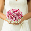 In this bridal close-up, the bouquet rests securely between the bride's gently curved hands, its rounded form a perfect sphere of blush-pink roses edged with delicate gypsophila, like a soft breath of cloud against her lace dress. The roses, each one fully unfurled, display beautifully layered petals in tones that range from pale shell pink at the outer edges to a richer blush at the centre, giving a painterly, almost watercolour effect. They are nestled so closely together that the eye moves in a smooth, uninterrupted circle around the bouquet, a classic design often requested by Canonbury couples seeking understated romance.

Tiny white baby's breath, or gypsophila, is threaded generously around and between the roses, its miniature blooms forming a fine, airy halo. The gypsophila softens the structure and echoes the delicate pattern of the bride's ivory lace bodice in the background, where fine floral motifs shimmer faintly in the gentle daylight. This interplay between rose petal and lace, between structured bloom and floating detail, creates a sense of lightness and grace, much like the calm, tree-lined streets running from Canonbury through to nearby Islington.

Though you cannot see them, you can almost imagine the stems bound neatly with ribbon beneath her fingers, keeping the bouquet compact and comfortable to hold during a long day of celebrations. The scent that rises from the flowers is likely sweet and romantic, with hints of green freshness underscoring the soft perfume of the roses. The atmosphere in the image is quiet but filled with emotion: a still moment before vows are spoken, captured in a bouquet that feels personal, tailored, and lovingly arranged by a local Canonbury florist who understands both tradition and the contemporary tastes of North London brides.