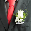 In the soft, diffused light the pale ivory rose of this boutonnière stands out against the smart charcoal jacket, its velvety petals layered in a quiet spiral that reads as both romance and resolve. Nestled at the rose's base, delicate sprays of baby's breath form a misty halo - tiny white orbs that temper the bloom's solidity with an airy purity. The wearer's checked shirt adds a muted texture behind the arrangement while a bold red silk tie introduces a touch of celebratory contrast, the deep crimson setting off the rose's cool tones with dramatic restraint. Slender slivers of fern and twisted leaves arch around the composition, their glossy surfaces catching light and imparting graceful movement; these green elements provide structure and a natural frame, making the boutonnière feel both botanical and architectural. A neat white ribbon bow at the stem supplies a final ceremonial detail, its satin sheen soft against the suit's wool. The fragrance is understated - a gentle rose note with a fresh, green undercurrent - conjuring images of early summer mornings along New River Walk or quiet walks through Canonbury Square gardens. Thoughtfully designed for a groom or special guest, this arrangement is an exercise in considered minimalism: tactile, cool, and composed, intended to be noticed in the small but meaningful gestures of a wedding day or milestone celebration. It embodies quiet confidence, crafted to sit perfectly against formal attire and to endure from ceremony through evening reception.